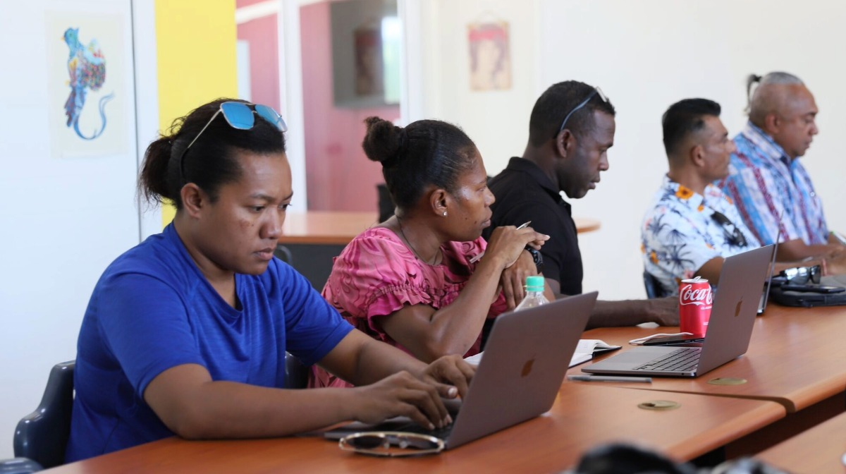 Five people sit at a table using laptops, focused on their work in a bright room.