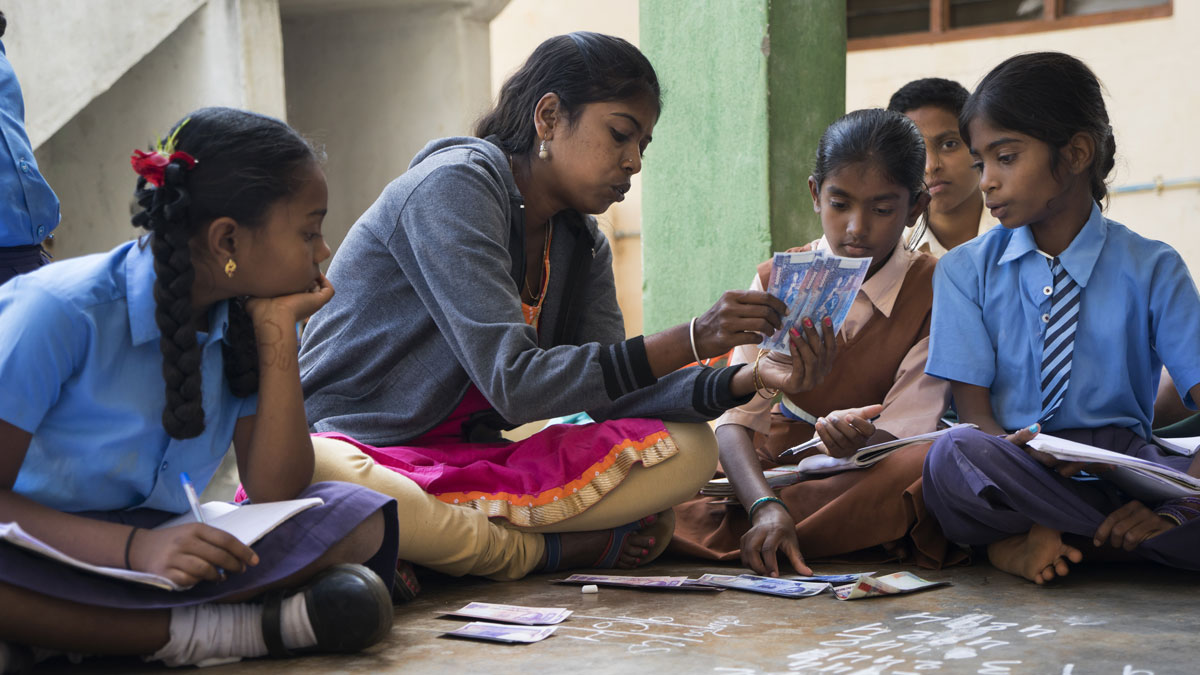 A teacher showing currency notes to students sitting on the floor in a classroom setting.
