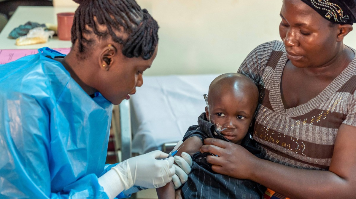 A healthcare worker in blue scrubs vaccinates a child held by a woman in a clinic setting.